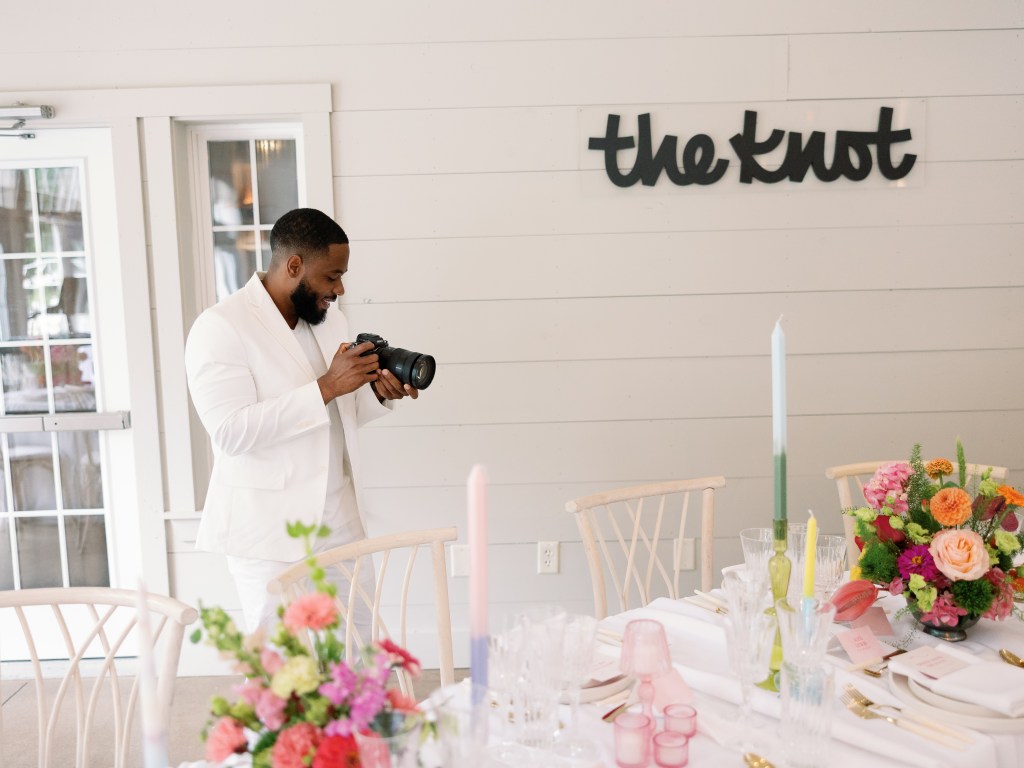 Man holding a camera at a wedding with The Knot logo behind him.